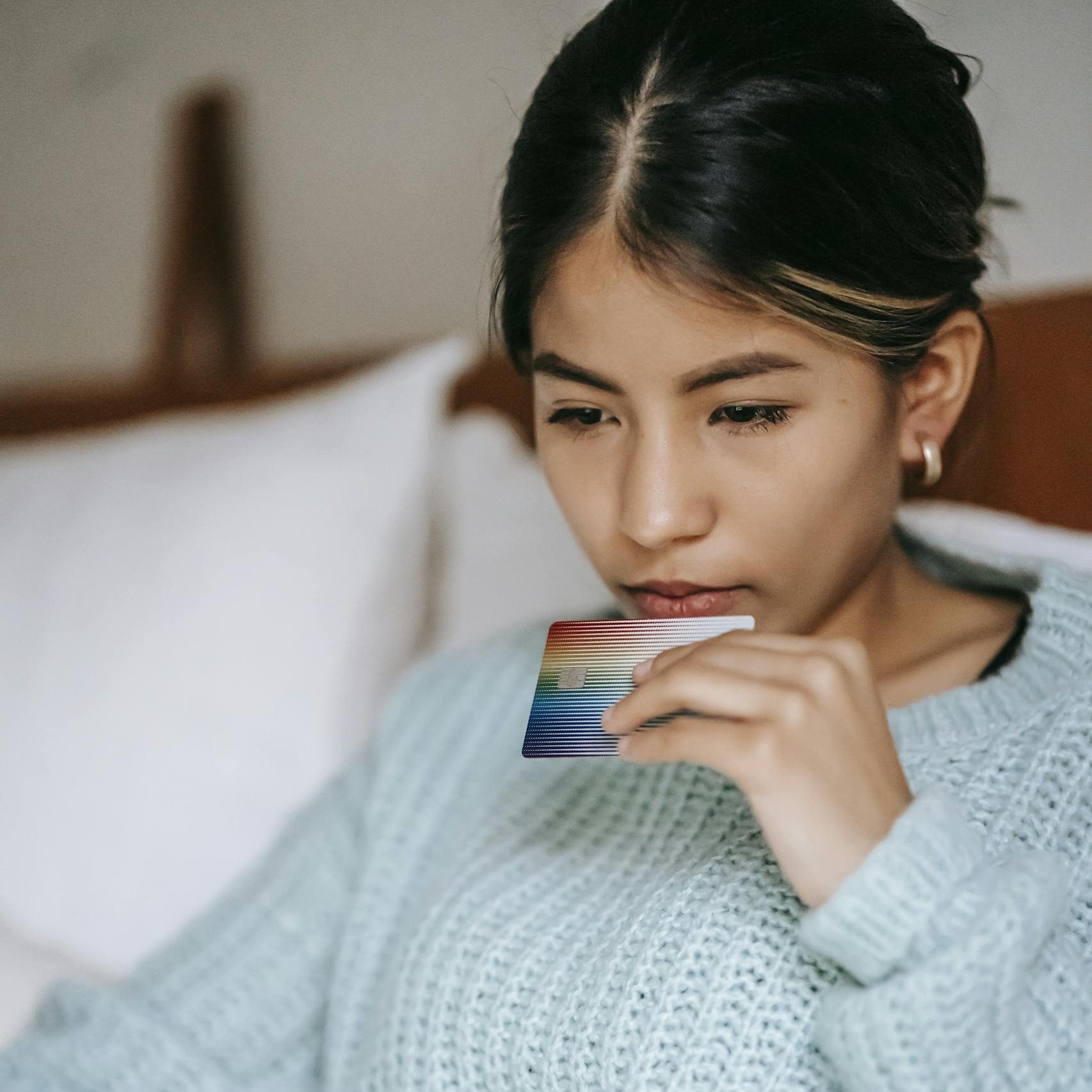 Crop concentrated Asian female wearing blue sweater making online payment while browsing netbook and lying on bed with credit card